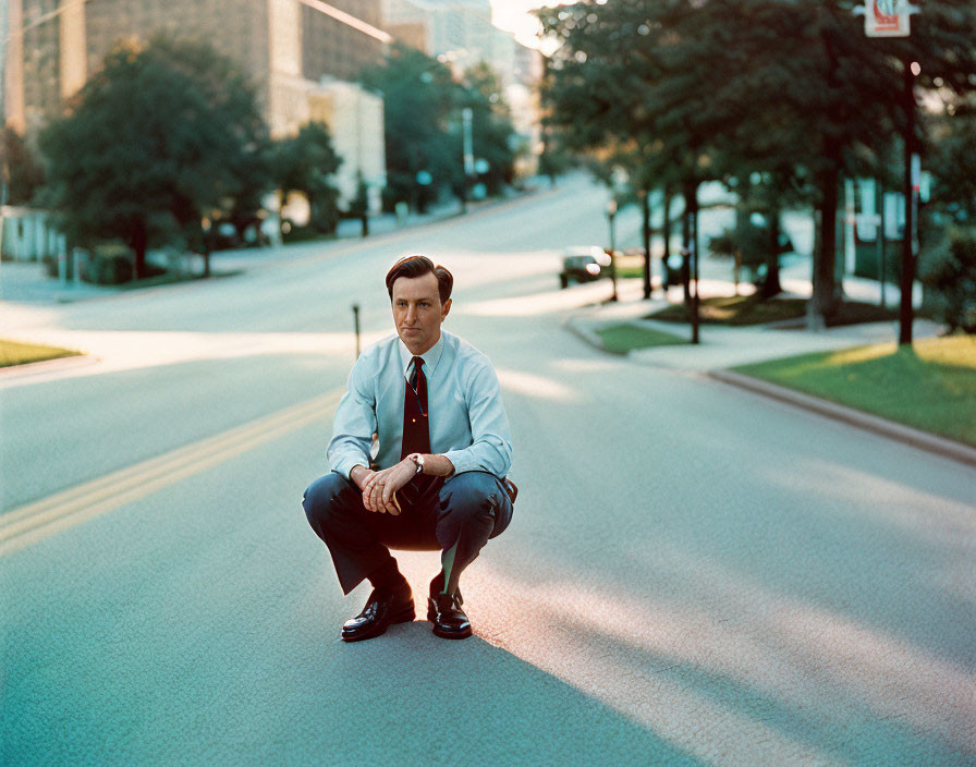 Man in suit squatting on empty street with trees and buildings in background