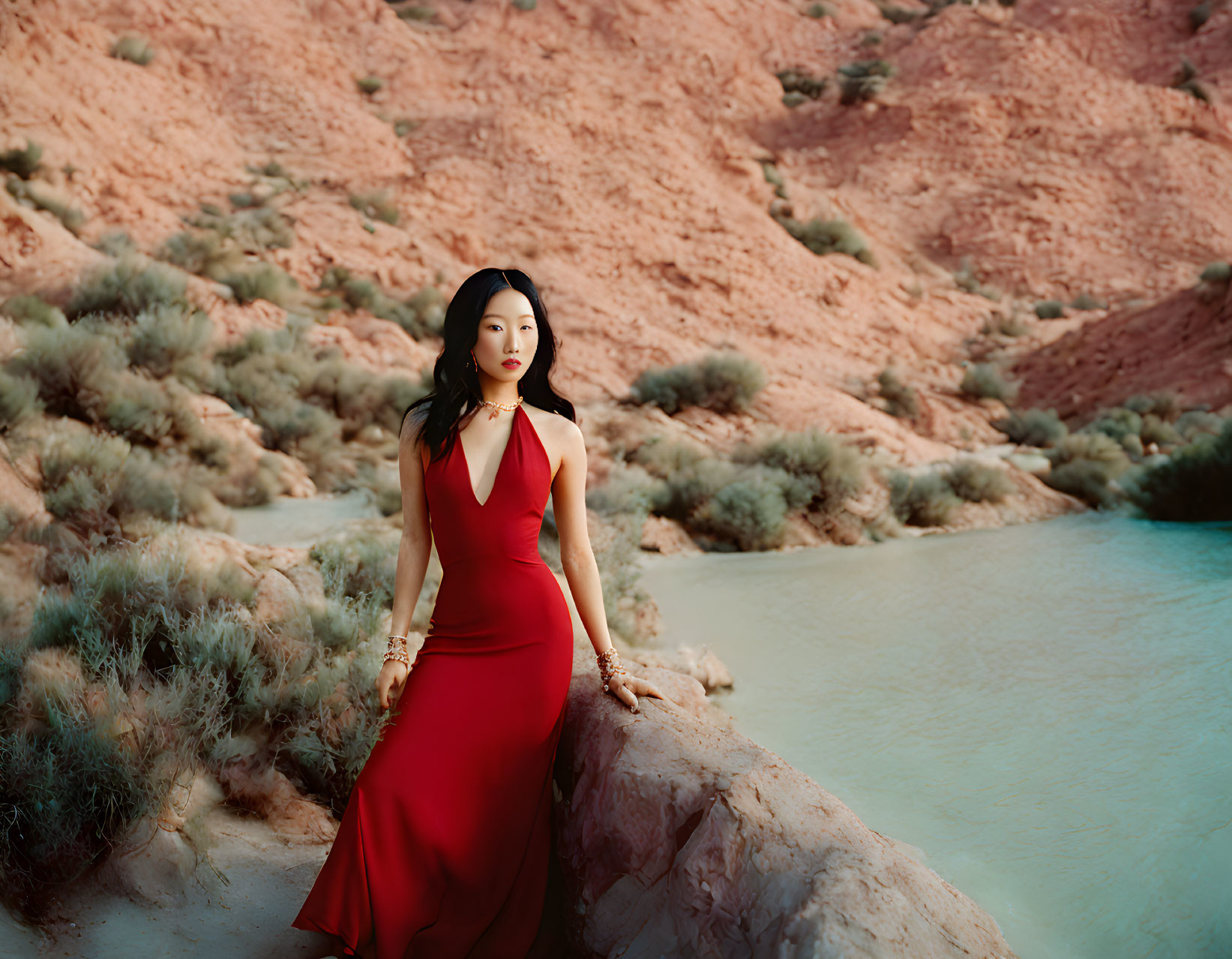 Woman in Red Dress by Blue Water Body and Red Rocky Hills