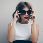 Short-haired woman in sunglasses holding shades, white top, looking sideways on light background