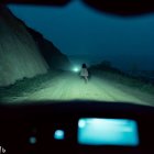 Person standing on dark road illuminated by headlights from car interior.