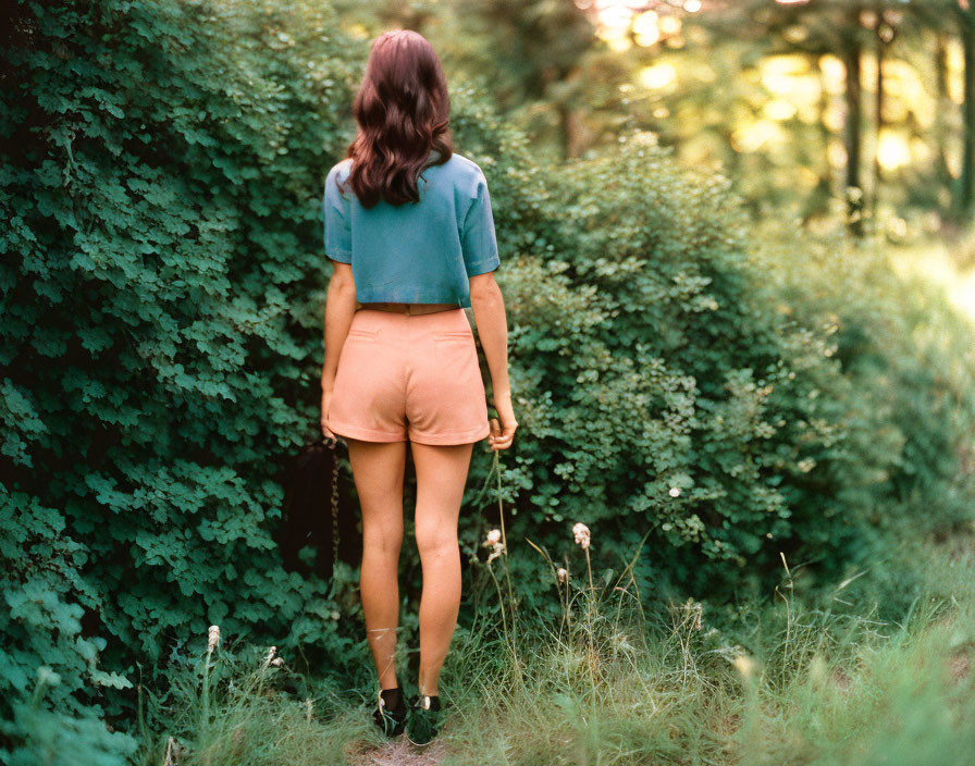 Dark-haired woman in teal blouse and orange shorts surrounded by lush greenery and wildflowers in forest.