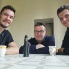 Three men at table with mugs, prosthetic arm, and debris.