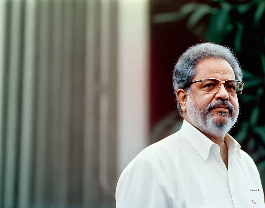 Elderly man with white hair and beard, glasses, white shirt, against vertical lines