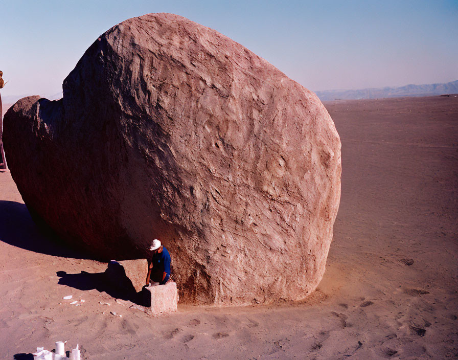 Person in hat next to massive boulder in desert landscape with blue skies and distant mountains