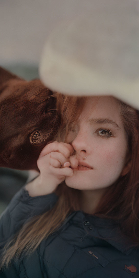 Red-haired woman in a pale hat gazes contemplatively.