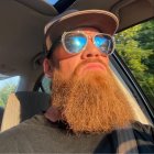 Bearded man in sunglasses and hat in car with tree reflections