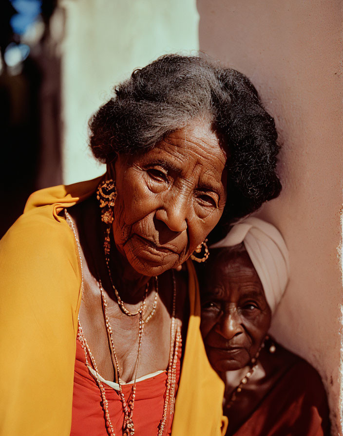 Elderly woman with gray hair and gold earrings in yellow top and red scarf, another woman in