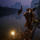 Person in hat and coat with lantern and bird by river at dusk with mountains