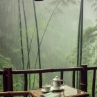 Balcony scene with bamboo chairs, cups, lantern, rain, and greenery