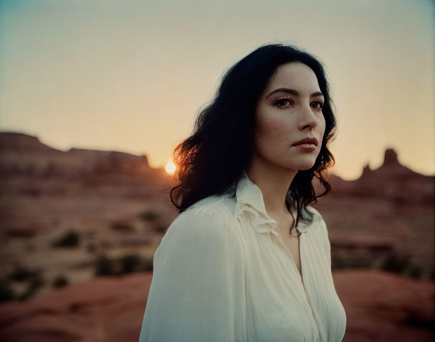 Woman in white blouse standing in desert at sunset with rock formations in background.