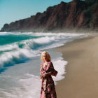 Woman in floral dress on sandy beach with waves and cliffs in misty background