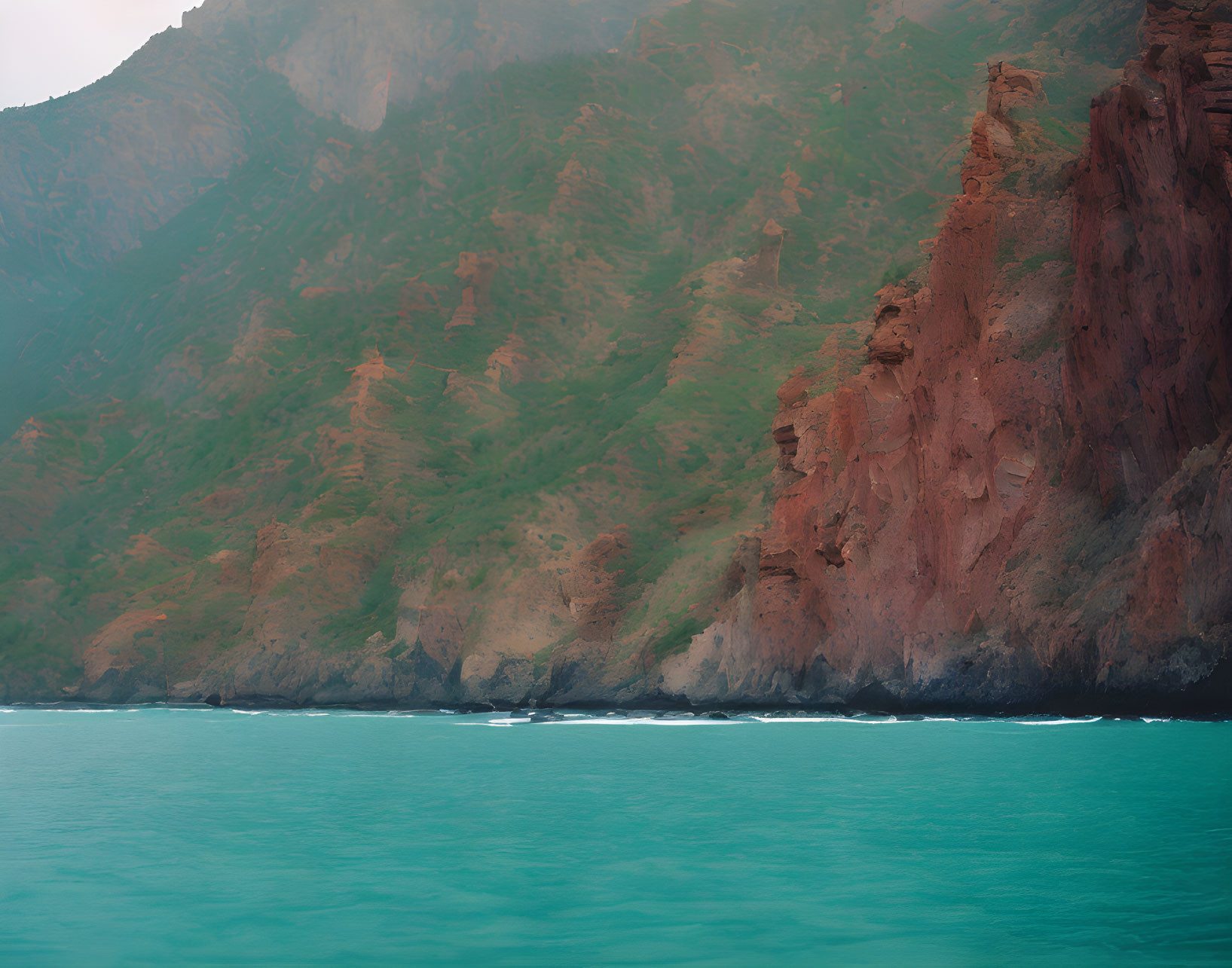 Scenic mountain slopes meeting turquoise sea with reddish cliffs and gentle waves.