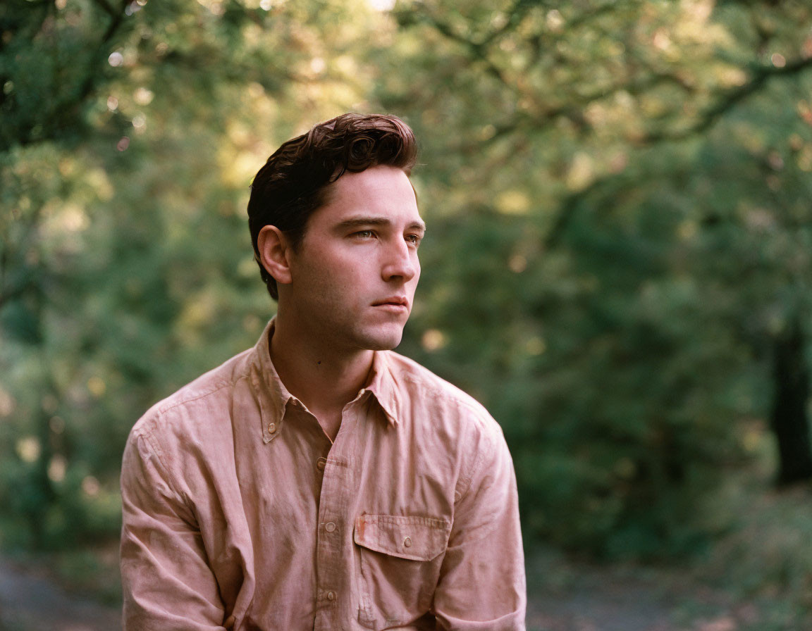 Young man in pink shirt sitting outdoors with green foliage background