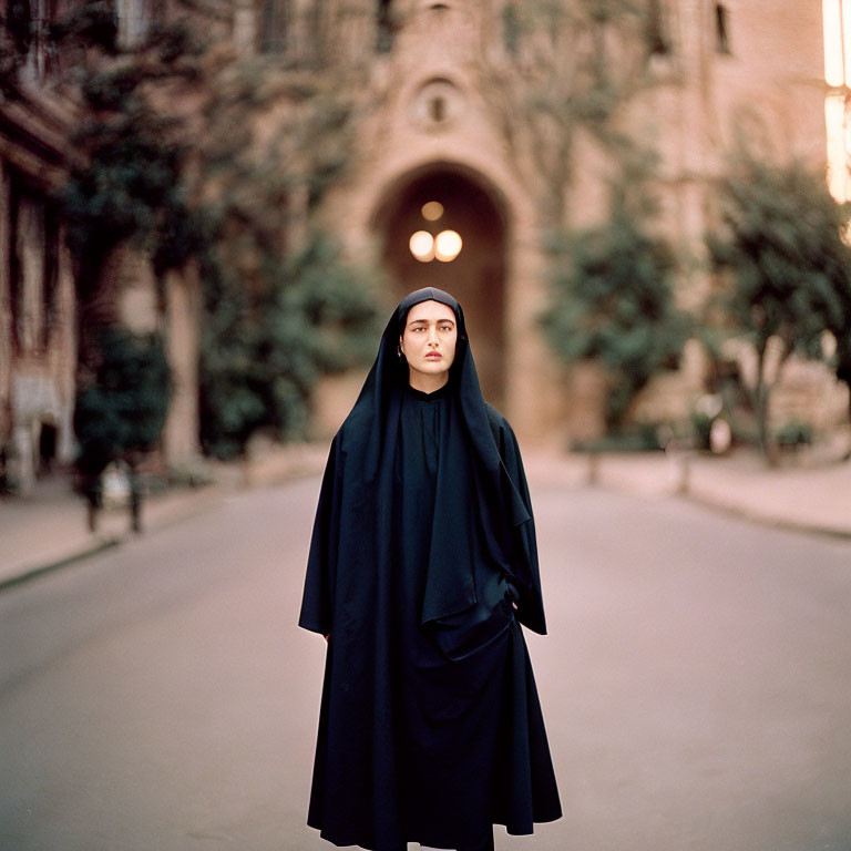 Woman in black cloak on path with archway, building, trees, soft-focus effect