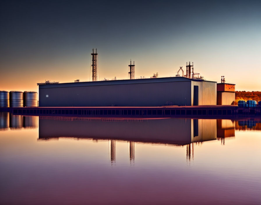 Industrial building reflected in calm water with golden sunrise or sunset, storage tanks on the left