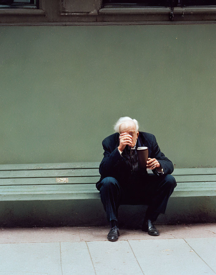 Elderly person with white hair sitting on bench in black suit with cane.