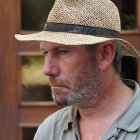 Weathered-faced elderly man in straw hat and blue shirt staring, rustic wooden structure in background