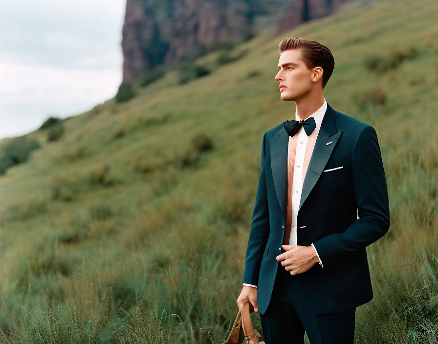 Man in formal black tuxedo with bow tie in grassy field with hills.