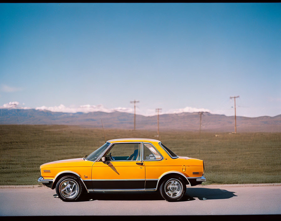 Vintage yellow car with black stripes and chrome accents parked by roadside with mountain backdrop.