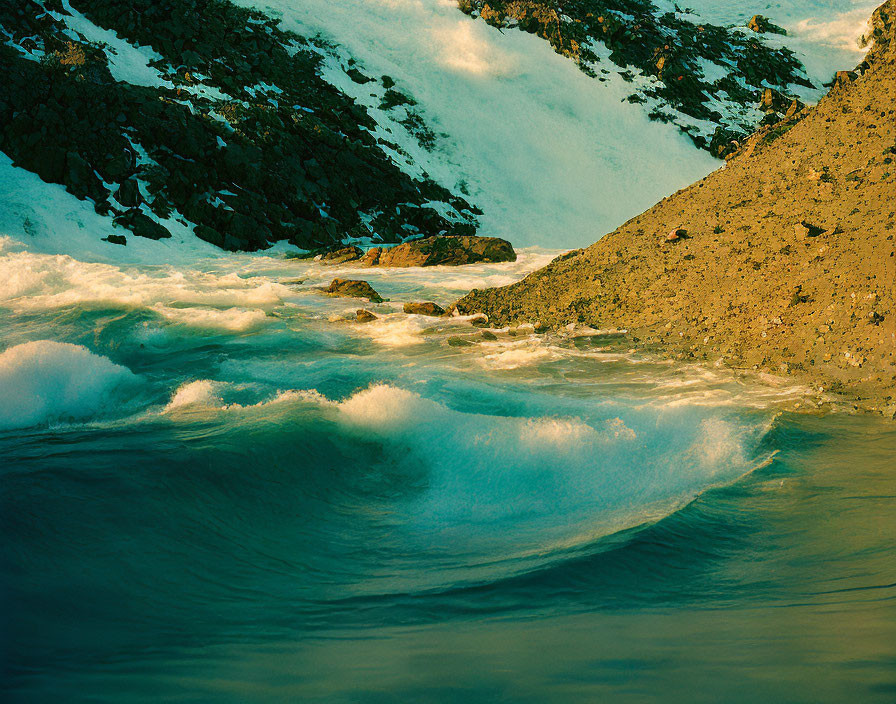 Rocky shore with snow-covered slopes and crashing waves in warm light