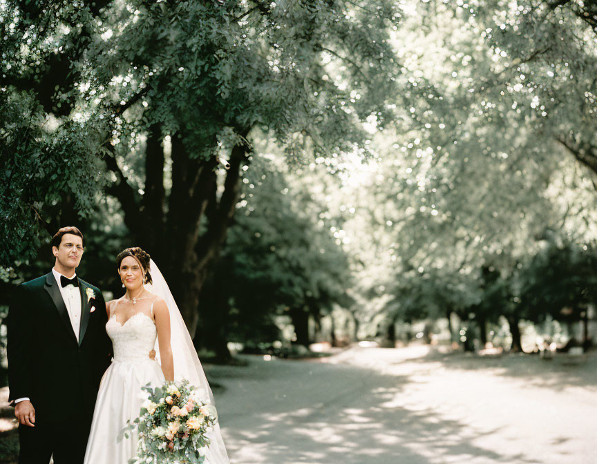 Bride and groom holding hands beneath tree canopy on sunlit road