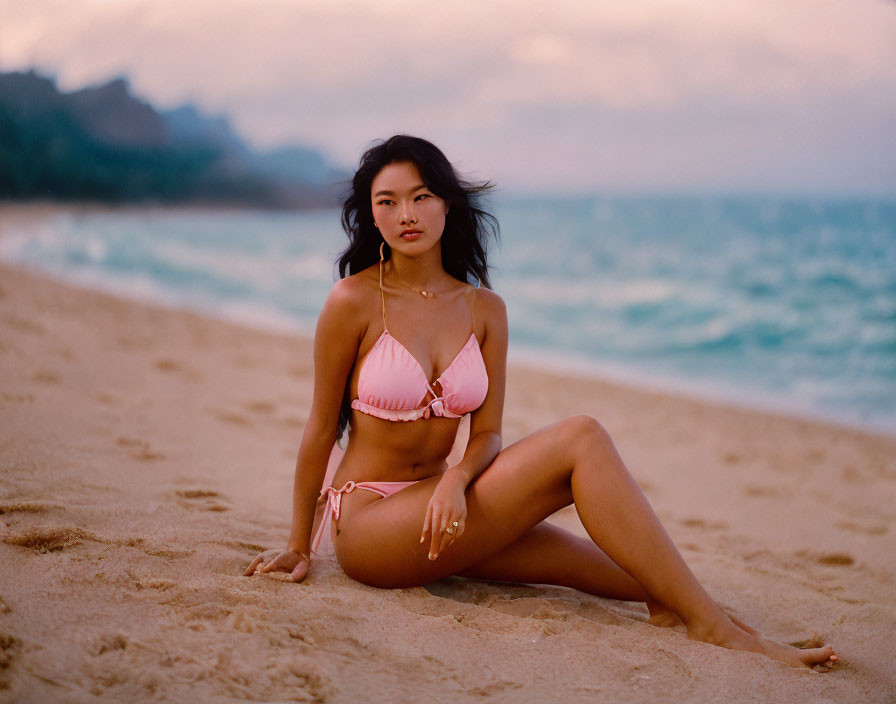 Person in pink bikini on sandy beach at dusk with ocean background.