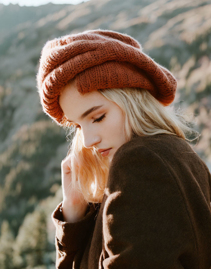 Contemplative woman in burnt orange knit hat against sunlit hills