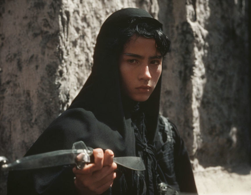 Young person with dark hair in black headscarf holding metallic object with focused gaze