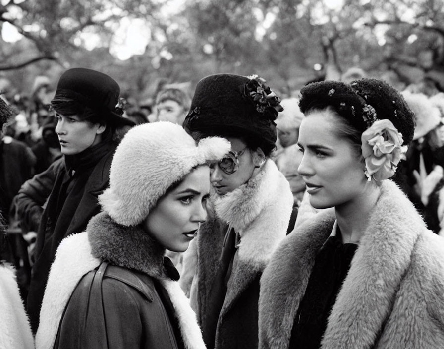 Vintage women in elegant attire and fur hats with a flower, black-and-white photo with crowd.