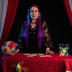 Colorful-haired woman at table with crystal ball, candles, and tarot cards in mystical setting.