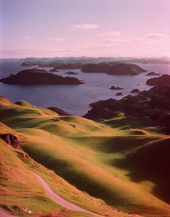 Tranquil sunset over green hills and serene sea with small islands.