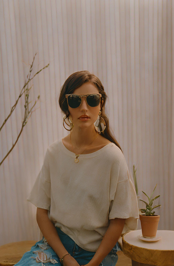 Woman in sunglasses and casual outfit sitting by corrugated wall with potted plant