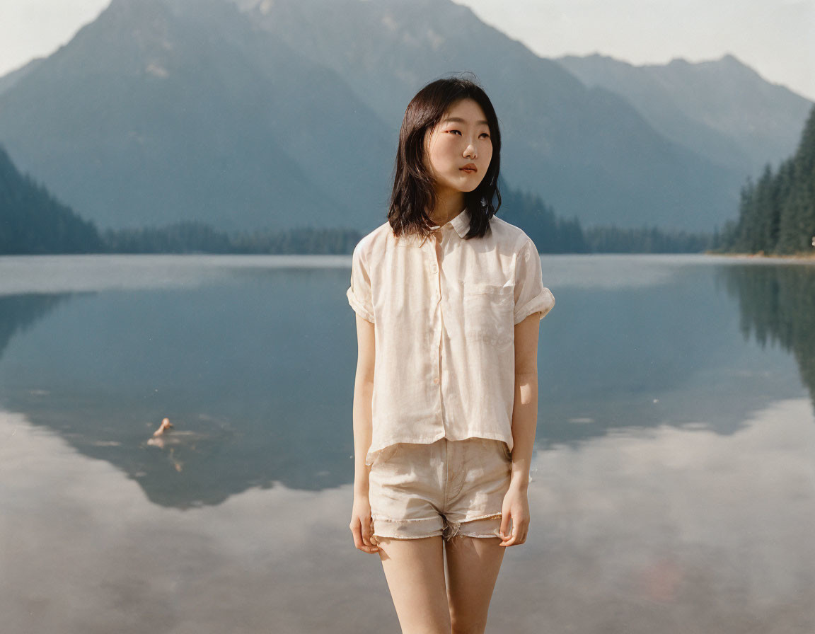 Woman in casual outfit by serene lake with forested mountains.