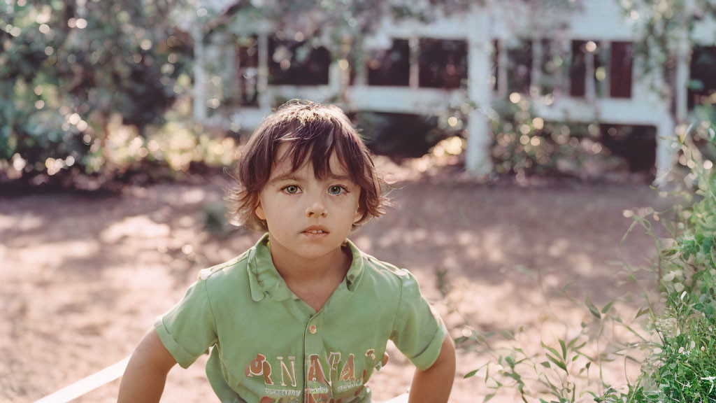 Child with tousled hair in green shirt outdoors with house and greenery.