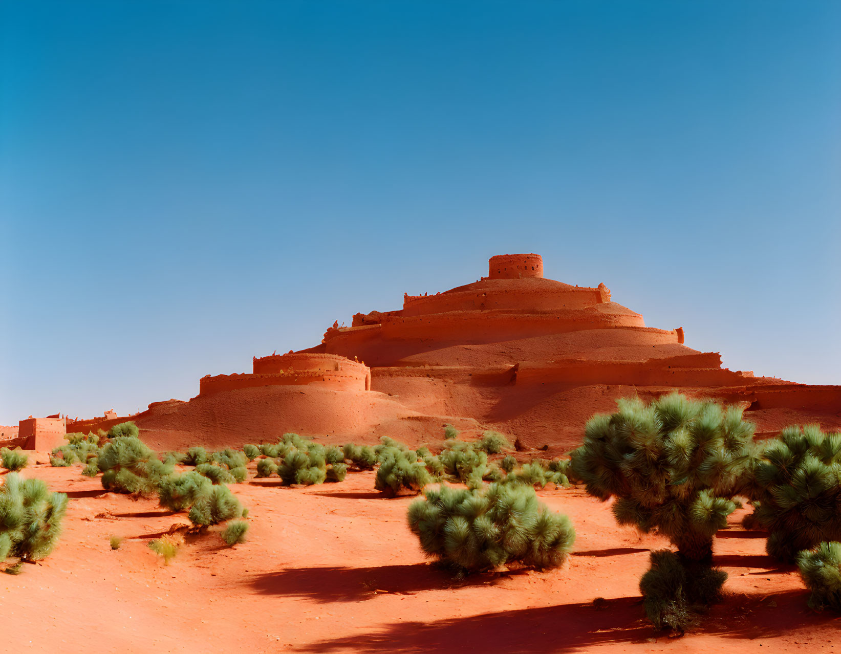 Ancient terracotta-colored stepped pyramid in Pueblo architecture setting on red sandy soil.