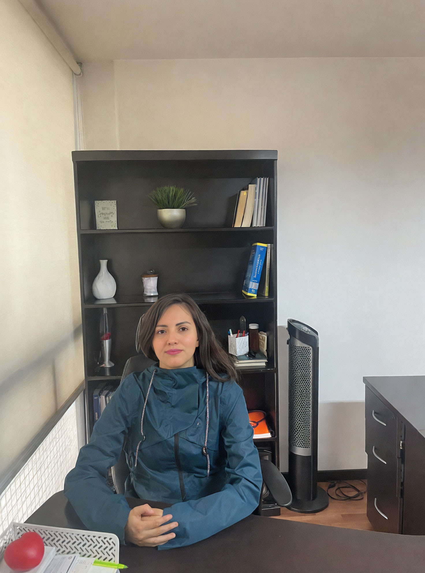 Woman in Blue Coat Sitting at Desk with Bookshelf and Decorative Items