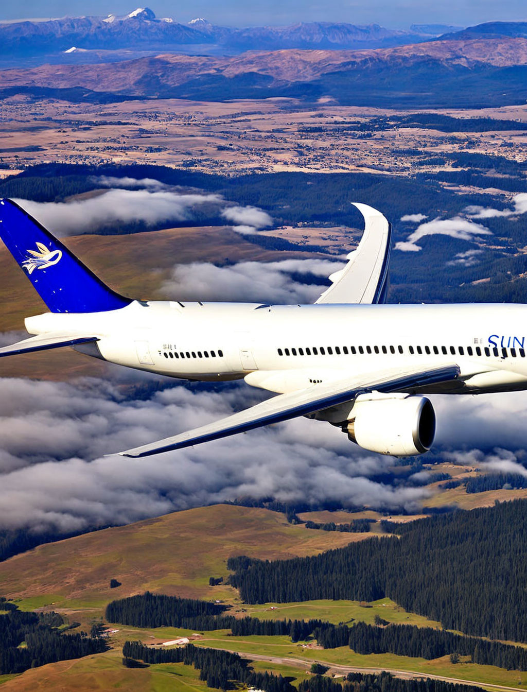 Commercial airplane flying over patchy clouds, rolling fields, and mountains.