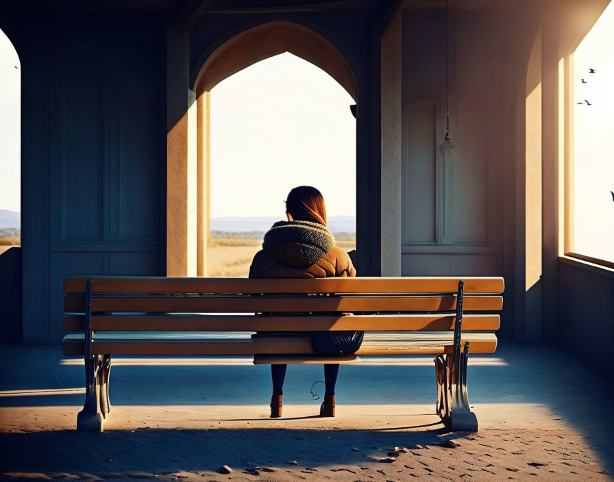 Person sitting on bench in shelter with arches, gazing at sunlit landscape