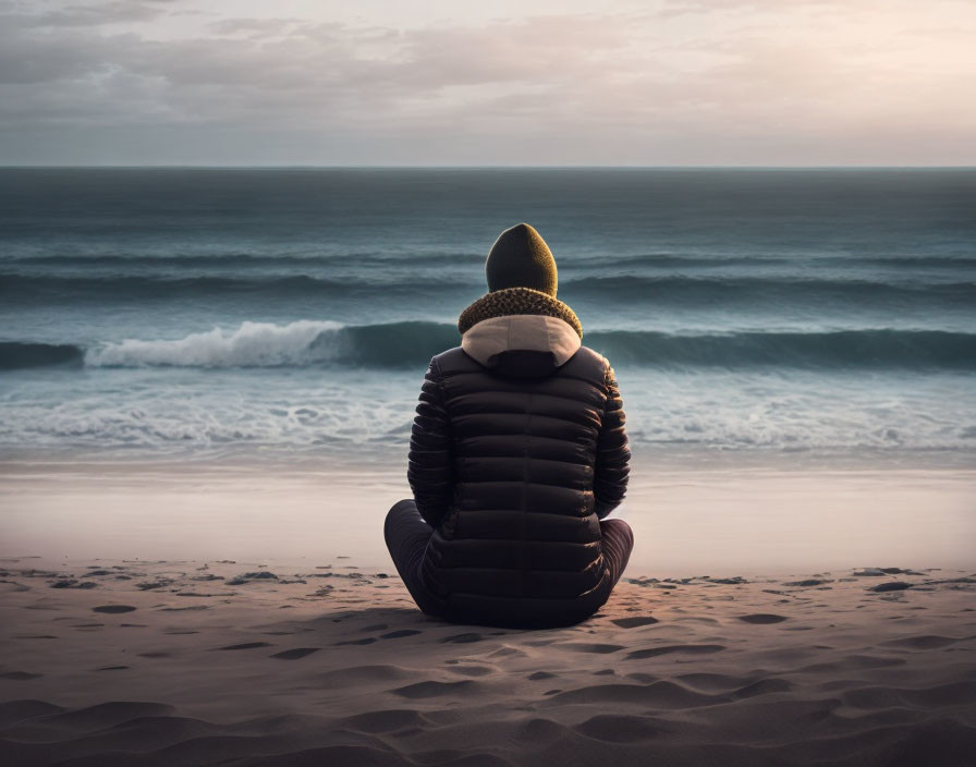 Person in warm jacket and beanie on sandy beach at dusk.