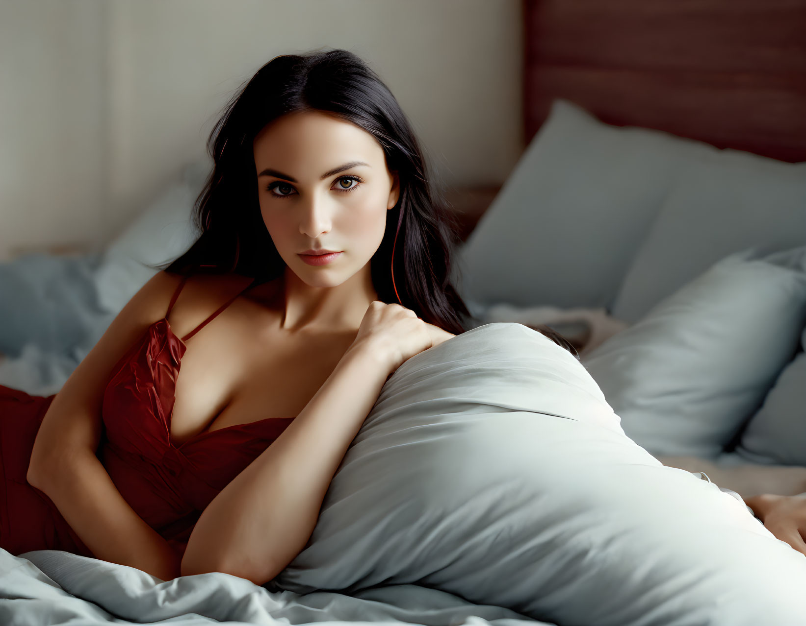 Dark-haired woman in red top reclining on bed with white pillows gazes at camera