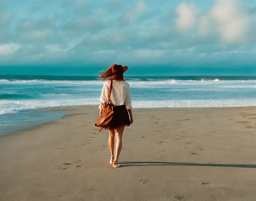 Woman in hat walking on sandy beach with blowing hair and skirt under cloudy sky