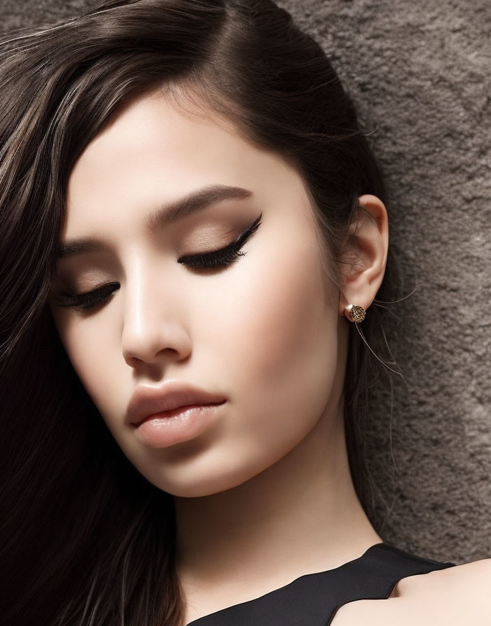 Portrait of woman with long hair, winged eyeliner, earring, leaning on textured wall.