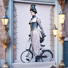 Vintage-style illustration of woman in period attire by bicycle with elegant street lamps and ornate blue doorway.