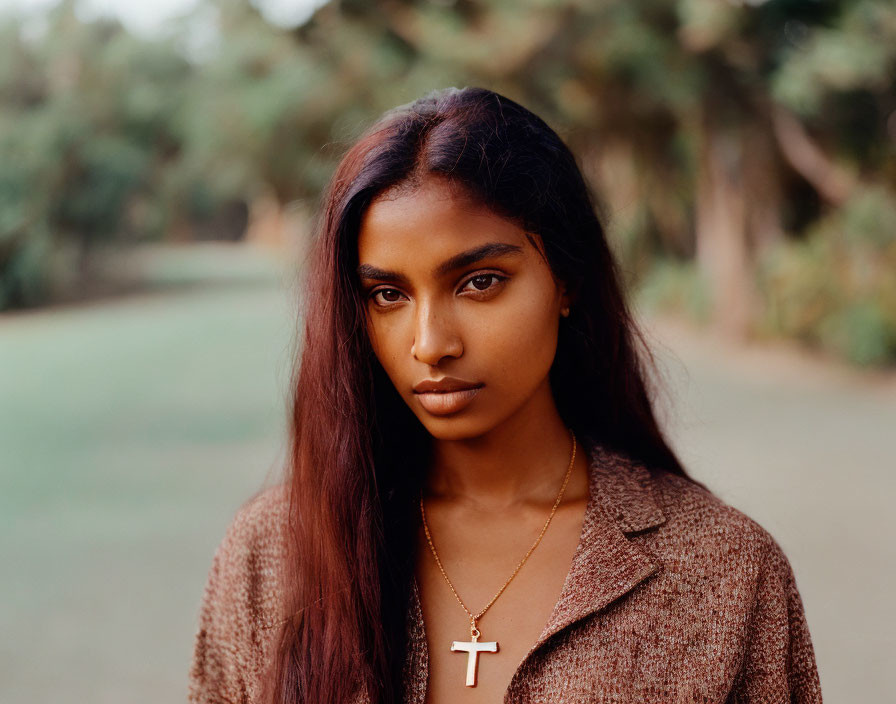 Portrait of woman with long hair, cross necklace, and brown jacket against blurred nature backdrop