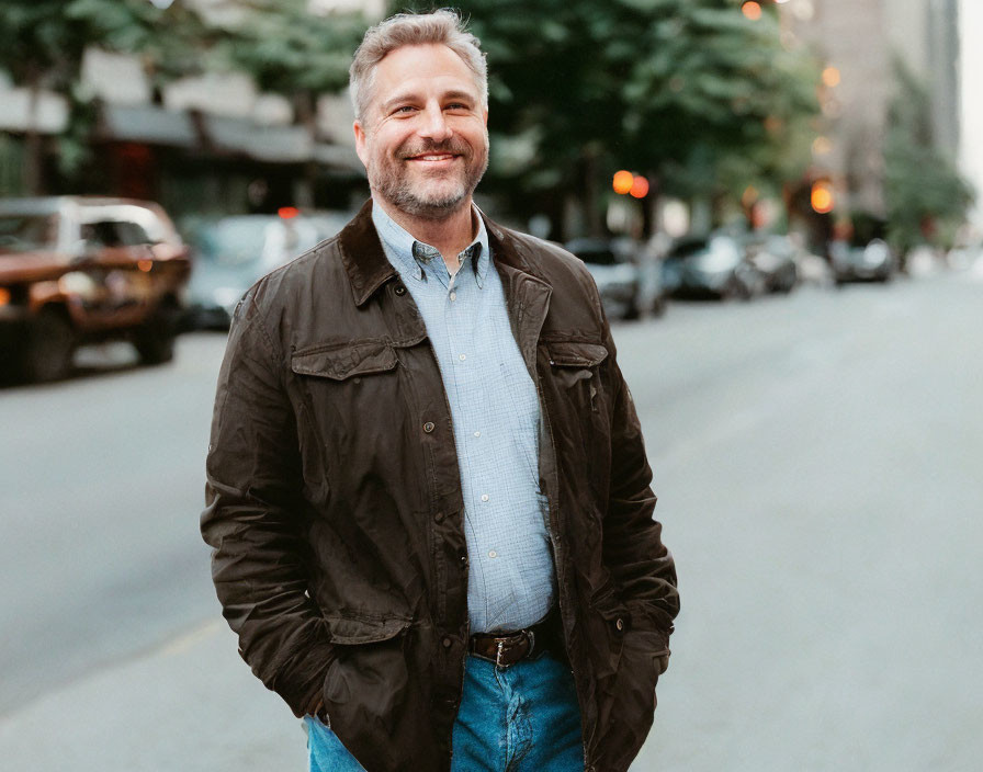 Smiling man with graying hair in brown jacket on city street