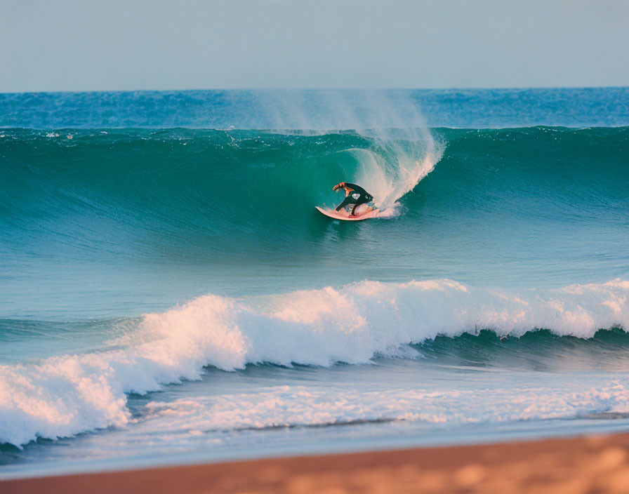 Surfer riding turquoise wave with spray against clear sky and sandy beach