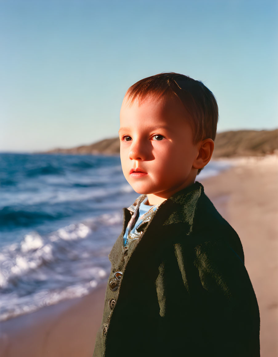 Young child in dark coat on beach with waves and clear sky