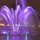 Colorful illuminated water fountain with purple lights against waterfall and greenery