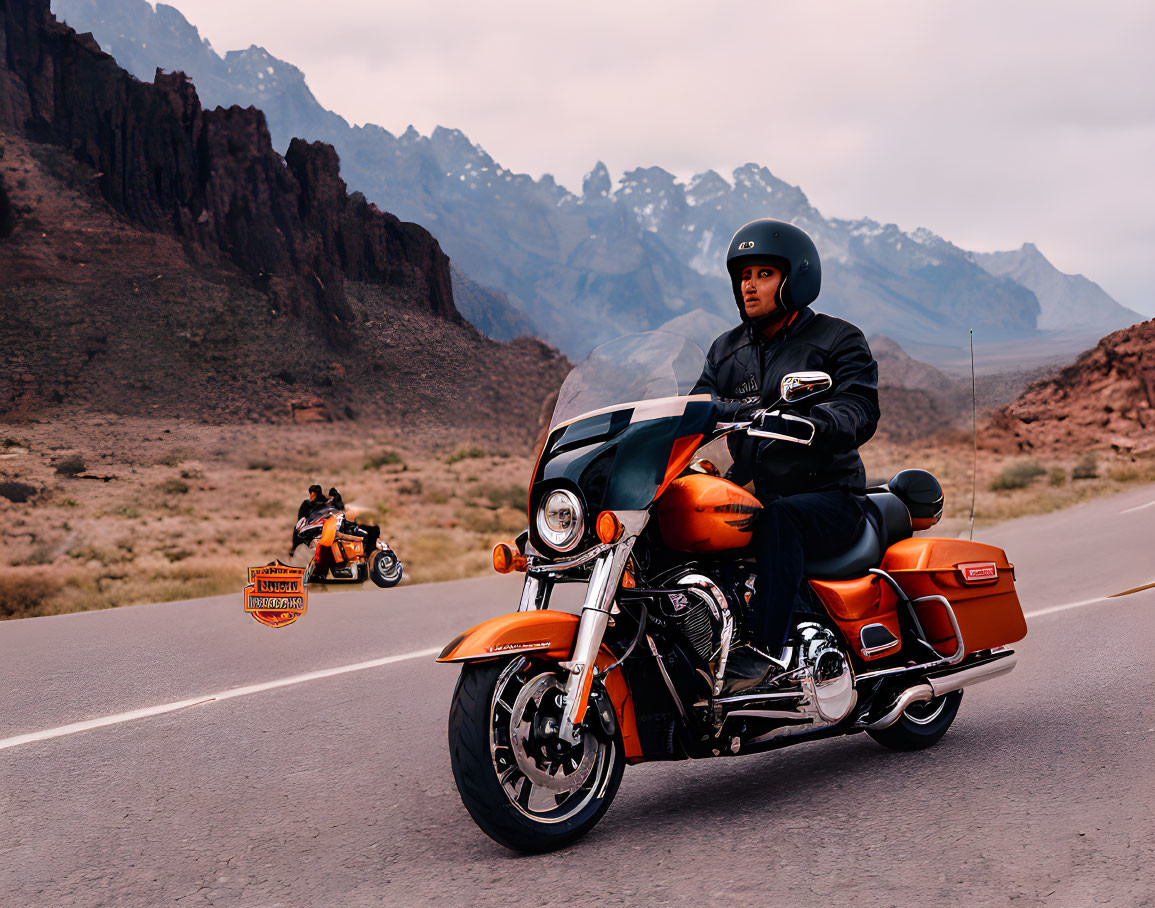 Motorcyclist in black gear on orange touring bike with mountain backdrop.