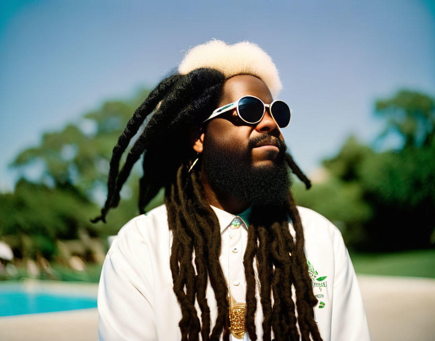 Man with dreadlocks and beard in sunglasses near pool under clear blue sky